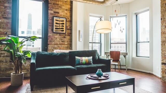 Modern living room with exposed brick walls, large windows, a dark green sofa, a coffee table, and a tall potted plant.