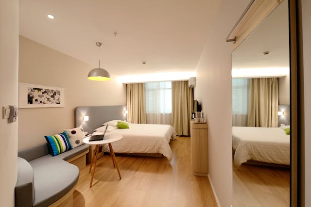 Modern, well-lit hotel room with two beds, a small round table, a couch with colorful pillows, and a wall-mounted TV.