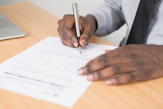 Person signing a document on a wooden desk with a silver pen, wearing a light shirt and dark tie.