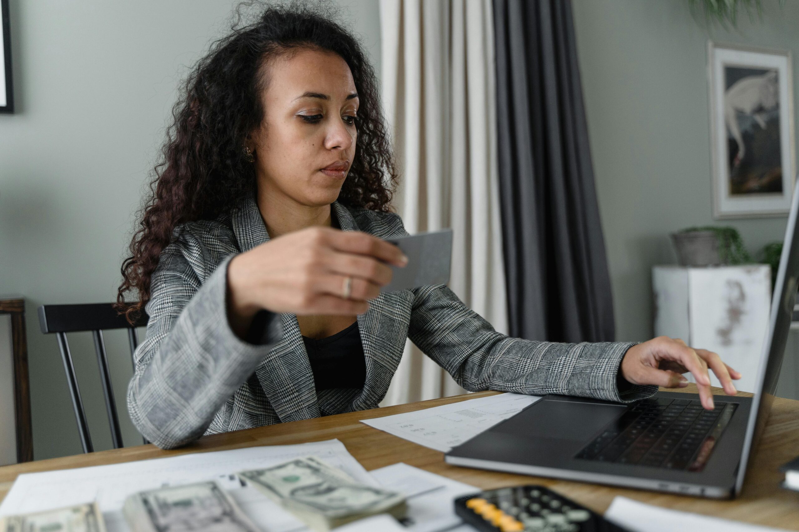 woman in a blazer sitting at a desk with an open laptop, cash, and a calculator
