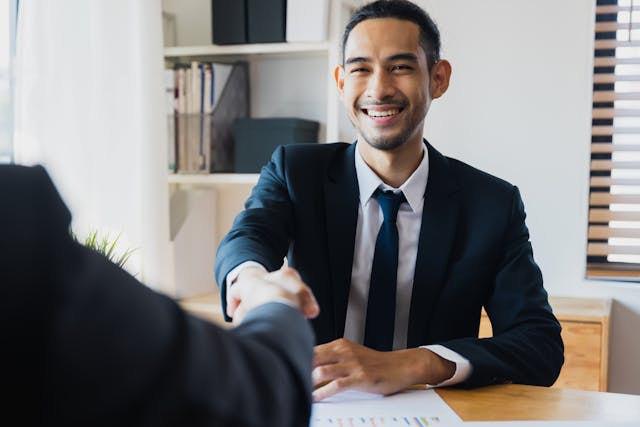a-smiling-man-in-a-suit-sits-at-a-desk-and-shakes-hands-with-another-person