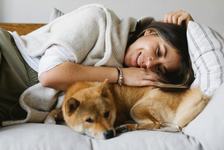 A smiling woman lying on a couch with her small reddish-brown dog.