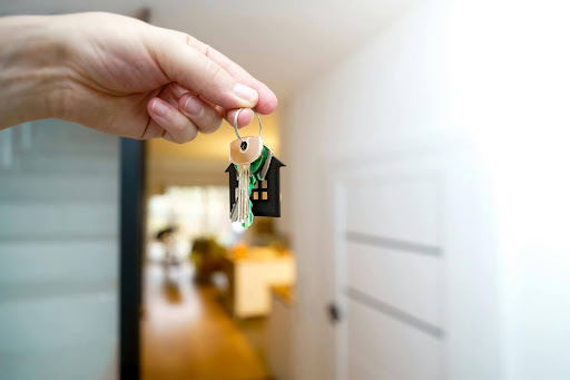 A hand holds a set of keys with a small house keychain, in a bright interior. 