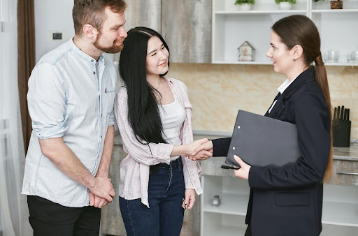 A couple smiles and shakes hands with a woman in a suit holding a folder.