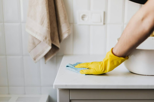 A person wearing a yellow glove wipes a white countertop with a blue cloth in a tiled kitchen