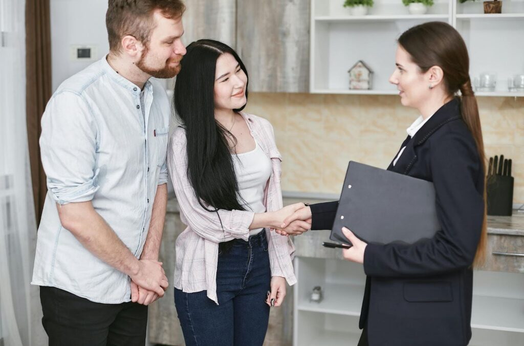 A smiling couple shakes hands with a woman in a suit in a modern kitchen.