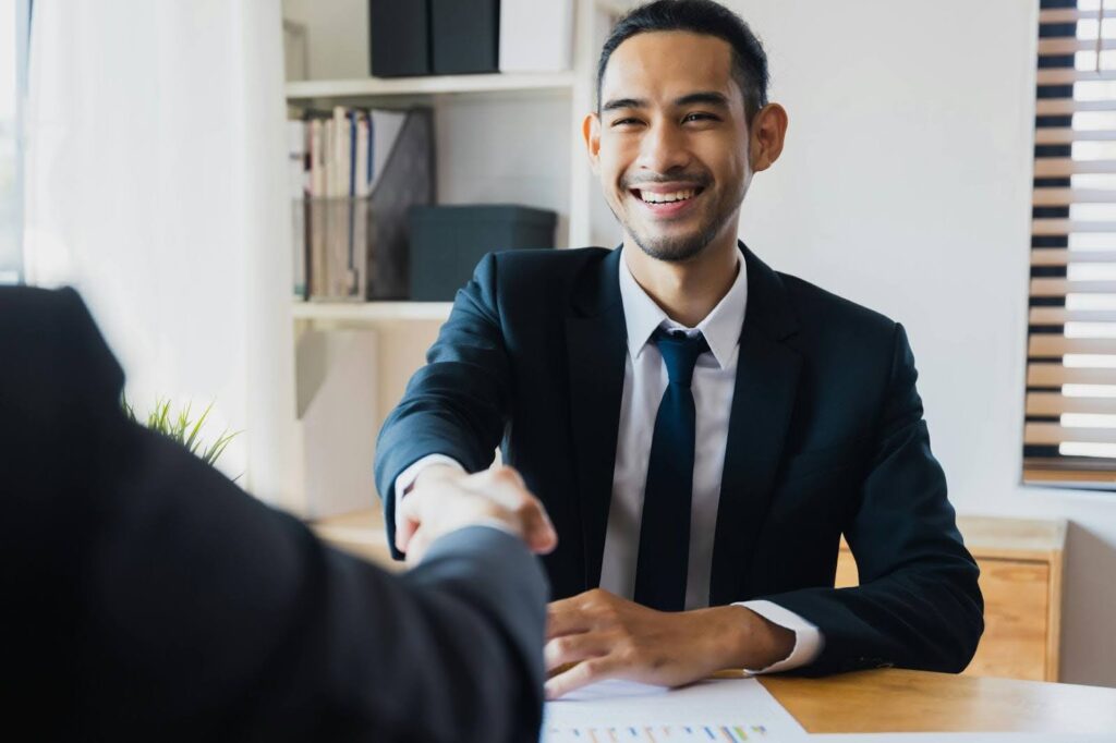 A man in a suit smiles warmly while shaking hands across a desk, suggesting a successful business meeting. Sunlight filters through a nearby window.