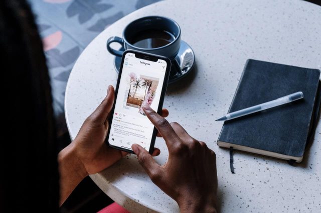 Person using a smartphone to browse Instagram at a round table, with a cup of coffee and a notebook with a pen beside them.